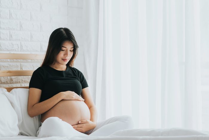 Happy Asian pregnant woman touching her belly with care in bedroom. She canâ??t wait to be mom.