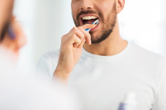 close up of young man with toothbrush cleaning teeth and looking to mirror at home bathroom