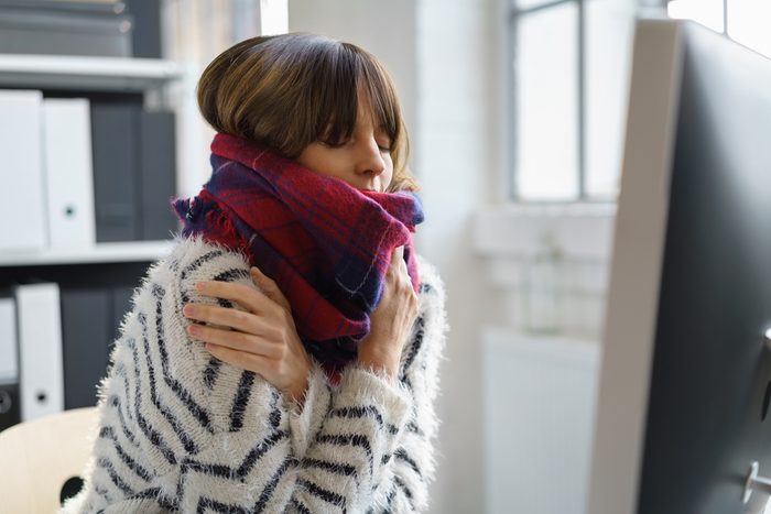 Sick businesswoman with winter chills and a fever sitting shivering in the office wrapped in a thick woolly winter scarf