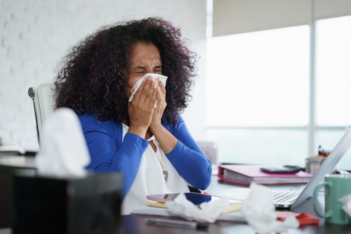 Sick african american girl working from home office. Ill young black woman with cold, sitting at desk with laptop computer and sneezing for allergy.