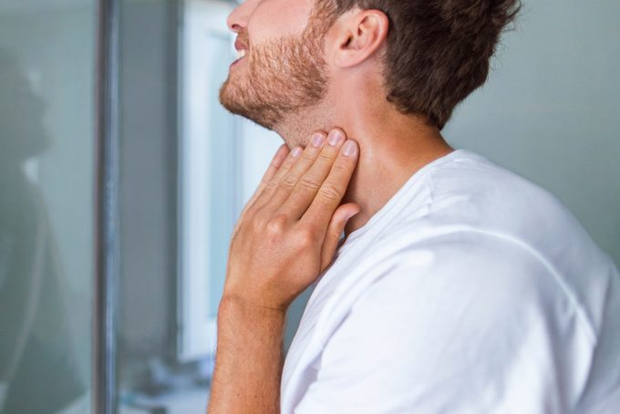 Thyroid self-exam checkup. Young man touching his neck at home bathroom doing self-check of his thyroid gland looking at mirror for early signs of health problem.
