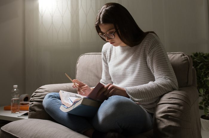 Young woman studying late at night at home, she is sitting on the armchair and reading a book, education and deadlines concept