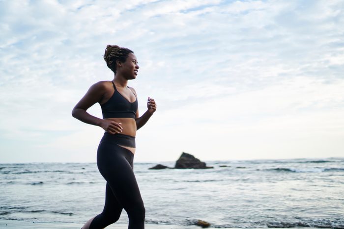 Side view of African American female jogger dressed in stylish sport clothes running along coastline washed by ocean water. Sportive fit woman with dark skin doing morning cardio training on seashore