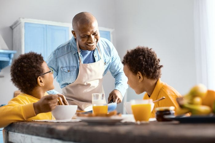 Happy family. Cheerful young man talking to his children and offering them omelet while they eating breakfast