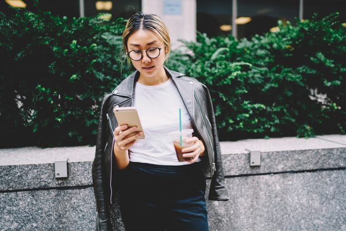 Young Chinese woman in glasses for eye protection reading news in website on smartphone connected to 4G internet. Female asian blogger chatting with followers in social networks while standing outside