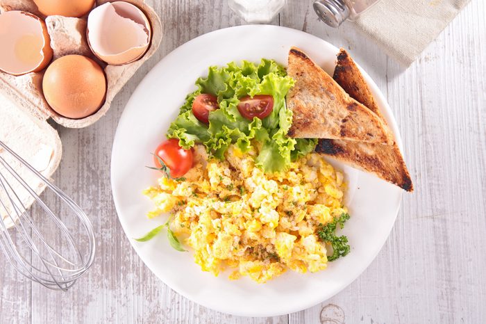 Breakfast meal of scrambled eggs, salad, and toast on a white table.