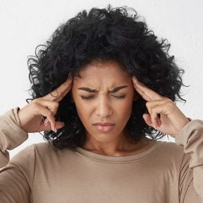 Dark-skinned young woman in beige long sleeved t-shirt frowning, having painful expressions, suffering from bad severe headache, keeping eyes closed, holding fingers on temples, trying to ease tension