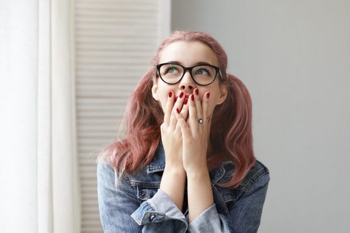 Isolated shot of fashionable young European woman with pink colored ponytails and red nails covering mouth with both hands and looking up with excited expression, overjoyed with great positive news