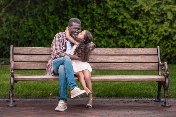 adorable african american girl hugging and kissing her grandfather while sitting on bench in park 