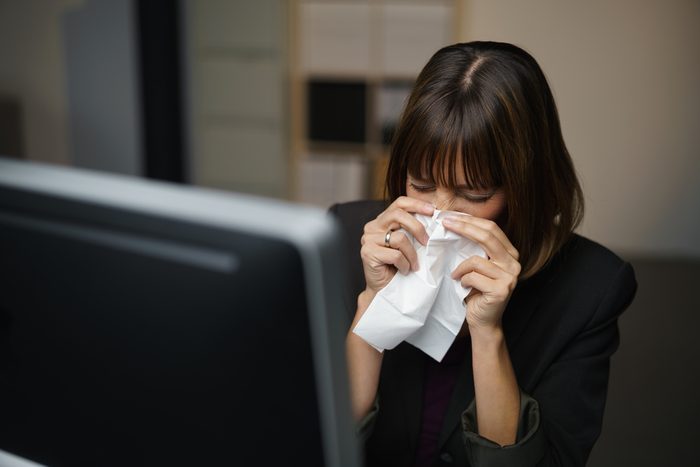 Woman sitting at a computer in an office, blowing her nose into a tissue
