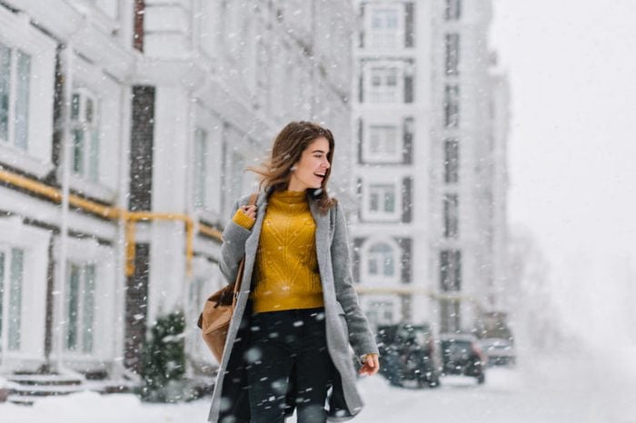 smiling young woman in coat with backpack walking in snowfall in Europe city centre. Expressing positivity, true emotions, enjoy snowing, waiting for christmas holidays, smiling to side