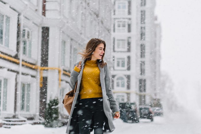 smiling young woman in coat with backpack walking in snowfall in Europe city centre. Expressing positivity, true emotions, enjoy snowing, waiting for christmas holidays, smiling to side