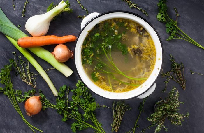 A large pot of homemade coocked fresh organic vegetable stock or broth with fennel, parsley, thyme, onions, carrot, mushrooms and leeks. Stone grey background with vegetables on it. 