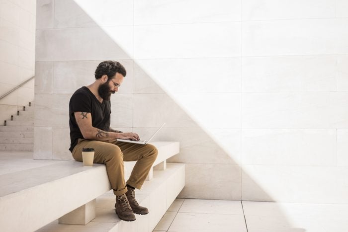Bearded man sits on shade of minimalistic and geometric space with his computer working on project
