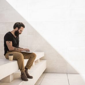 Bearded man sits on shade of minimalistic and geometric space with his computer working on project. Man is very concetrated and wears generic stylish outfit with leather boots. Space for your text.