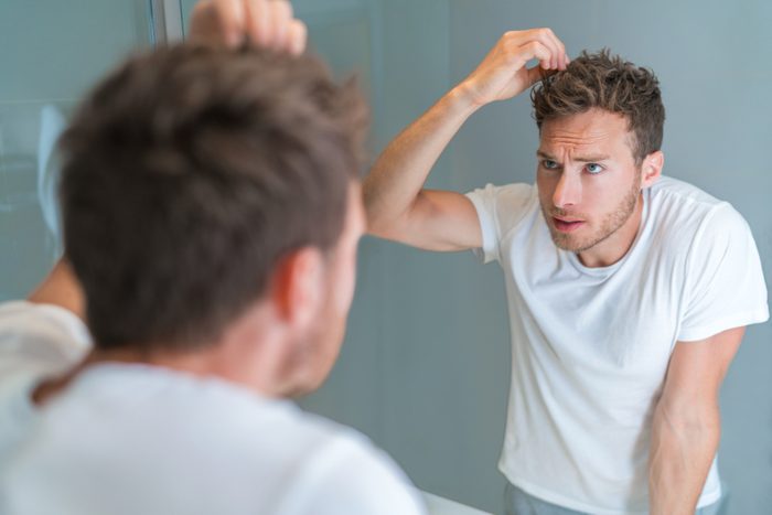 Hair loss man looking in bathroom mirror styling hairstyle with gel or checking for hair loss or grey hairs. Unhappy male health problem.