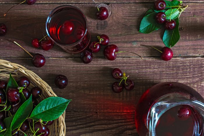cold cherry juice in a glass and pitcher with cherries inside on wooden table with ripe berries in wicker basket. top view