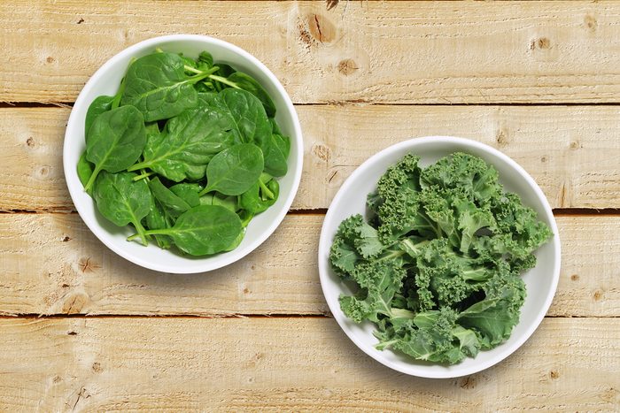 Two white bowls one containing spinach leaves and one containing chopped kale leaves