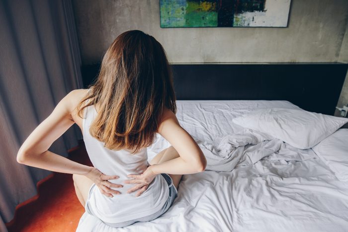 Young woman sitting on the bed holding her back with a backache.