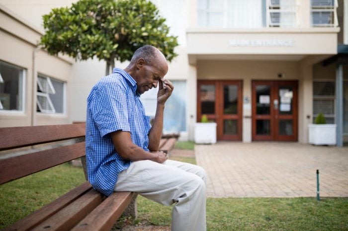 Sad senior man sitting on wooden bench outside nursing home