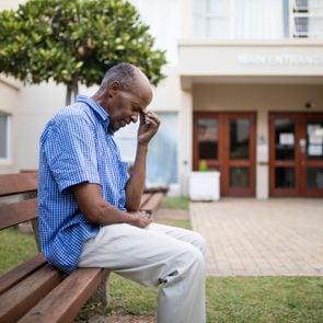 Sad senior man sitting on wooden bench outside nursing home