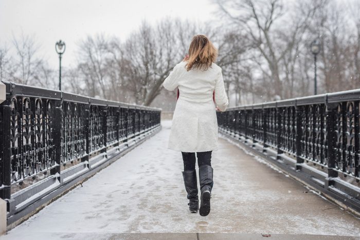 one woman walking on bridge in city park with snow falling and back to camera