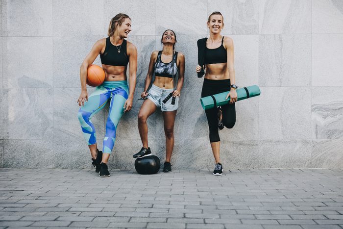 Happy fitness women standing outdoors against a wall holding basketball skipping rope and exercise mat. Fitness women having fun standing on street after workout.