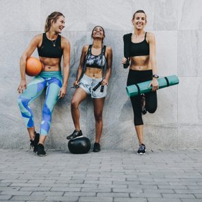 Happy fitness women standing outdoors against a wall holding basketball skipping rope and exercise mat. Fitness women having fun standing on street after workout.