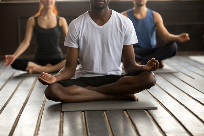 Young black man and a group of sporty people practicing yoga lesson with instructor, sitting in Sukhasana exercise, Easy Seat pose with mudra gesture, working out, indoor close up, studio floor