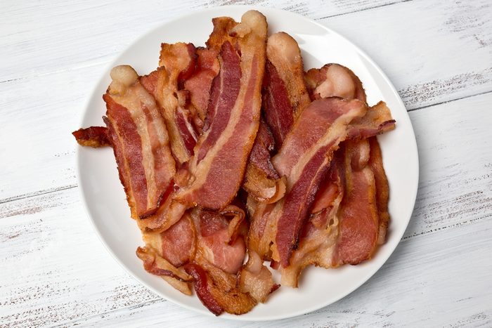 A small white plate of fried bacon strips on a white painted background.