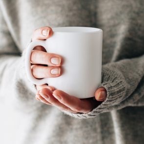 Closeup of female hands with a mug of beverage. Beautiful girl in grey sweater holding cup of tea or coffee in the morning sunlight. Mug for your design. Empty.