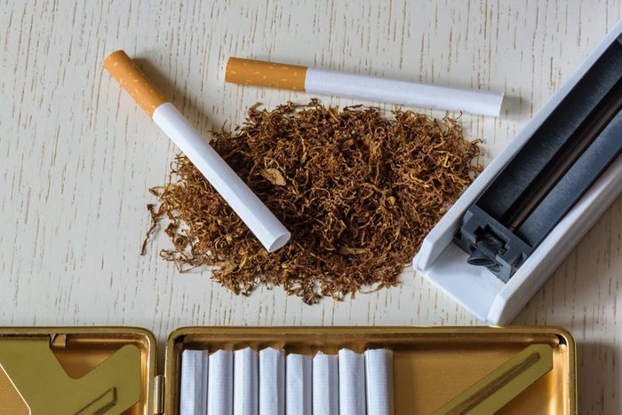 A pile of natural tobacco on a white wooden table, a device for manual production of cigarettes and cigarettes in a cigarette case. Smoking. Nicotine addiction. Health hazard.
