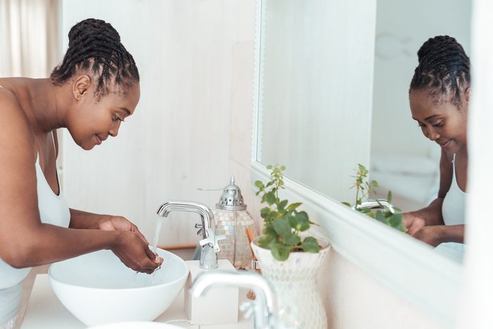 Young African woman standing over the sink in her bathroom washing her face with water in the morning