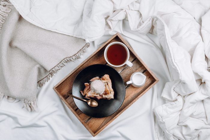 Cozy flatlay of bed with wooden tray with vegan apple pie, ice cream and black tea on white sheets and blankets