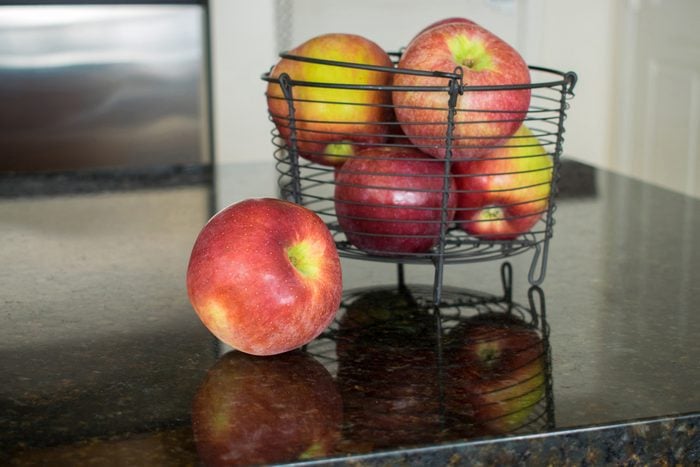 One apple beside a metal basket filled with red apples and strong reflections.