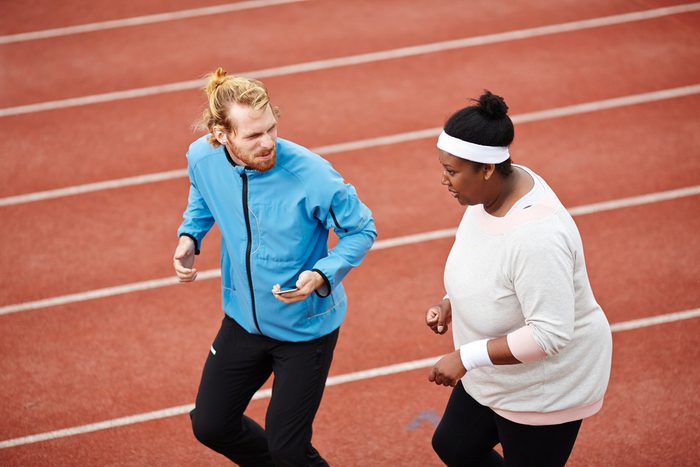 Young instructor and plump female jogging together on stadium racetrack before marathon competition