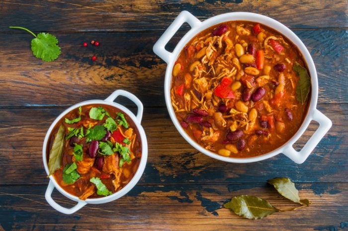 Turkey chili, ,stewed with beans, tomatoes, bell pepper, onion, garlic, thyme, cinnamon, chocolate and fresh cilantro, in white bowl and casserole on wooden table.