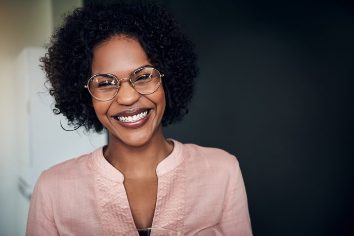 Young African businesswoman wearing glasses and laughing while standing alone in a modern office