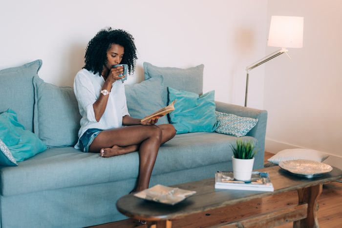 Beautiful young black woman seating in the sofa reading a book and drinking coffee