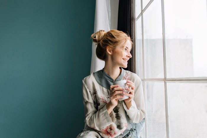Portrait of young woman with blonde hair drinking coffee or tea next to big window, smiling, enjoying happy morning at home. Turquoise wall on background. Wearing silk pajamas in flowers.