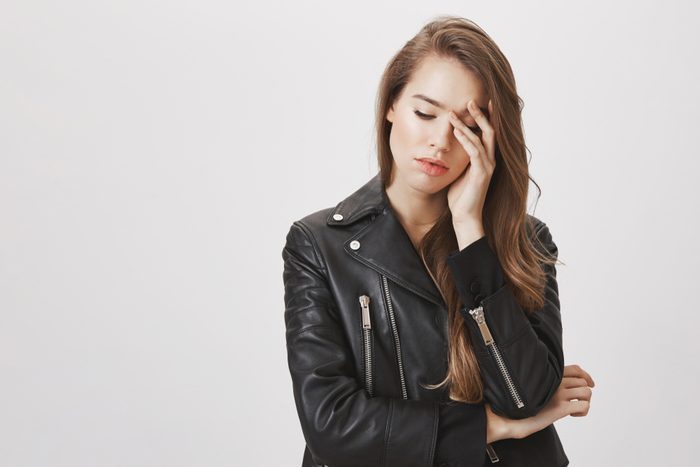 Studio shot of gloomy feminine urban woman in leather jacket looking down and touching forehead with hands, feeling miserable or exhausted, having headache from stress, standing over gray background