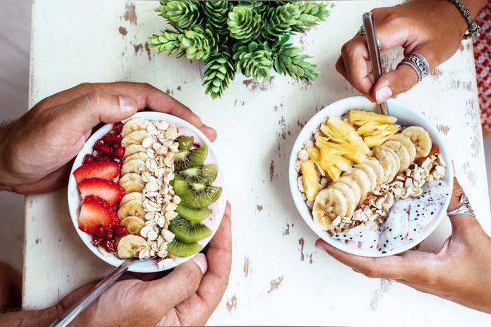 Smoothie bowls with mixed tropical fresh fruits on wooden table with hands, top view from above. Summer healthy diet, vegan breakfast.