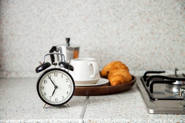 Breakfast concept. The alarm clock, coffee, croissant in an interior of kitchen. Selective focus