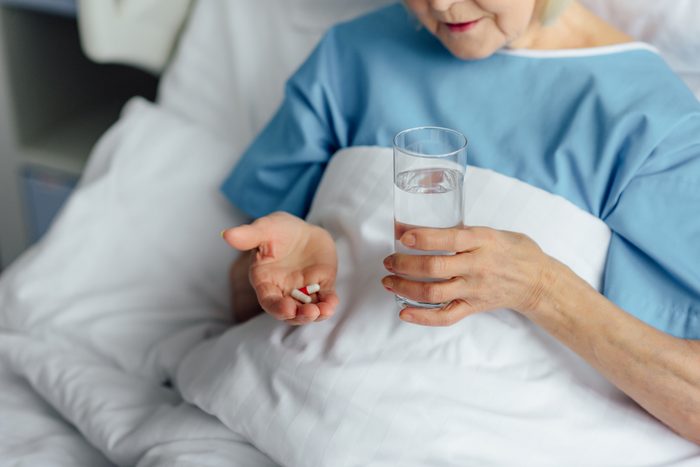 cropped view of senior woman lying in bed and holding pills with glass of water in hospital
