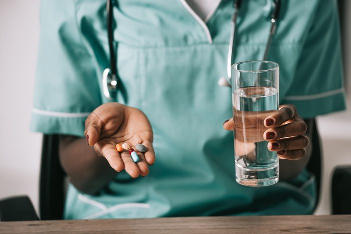 cropped view of african american nurse with stethoscope holding glass of water and pills in hand
