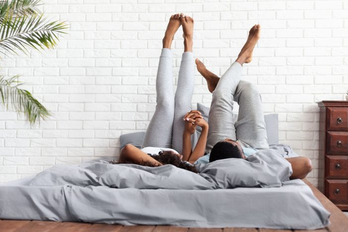 Couple in love. Young happy african-american man and woman lying on bed at home raising legs up, lazy family resting on weekend, copy space