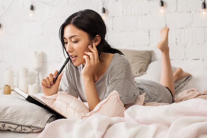 Young asian girl lying in bed, relaxing in her bedroom and write notes. Asian female student doing homework in bed. Girl in casual dress writing diary or studying in bed thinking.