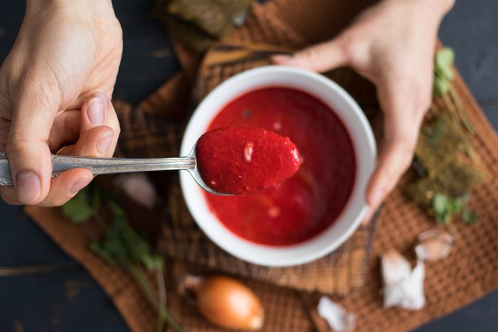 Young woman's hands holds spoon and bowl of traditional Russian beetroot soup - borscht. Raw and vegan with tomatoes, avocado, walnuts, parsley, garlic, onion. Vegetarian healthy food. Top view