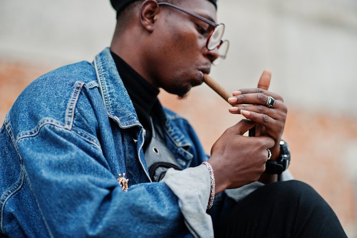 Black man in a denim jacket, beret, and eyeglasses lights a cigar.