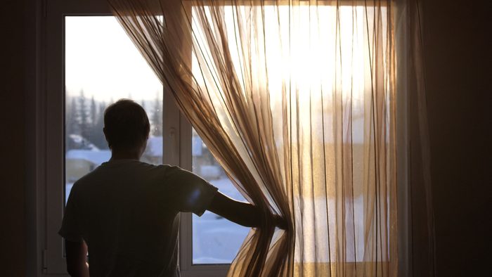 Young male opens curtains to bright sunny winter day and looks out of window through the sun during sunrise